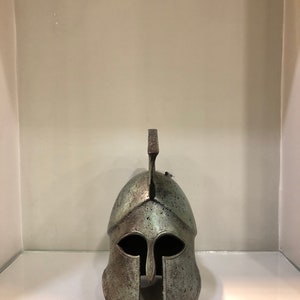 May include: A bronze Greek warrior helmet with a plume on top. The helmet has a face mask with eye holes and a nose guard.