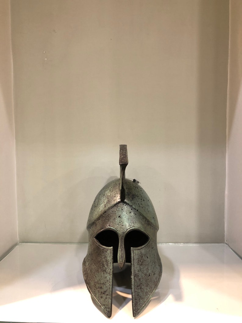 May include: A bronze Greek warrior helmet with a plume on top. The helmet has a face mask with eye holes and a nose guard.