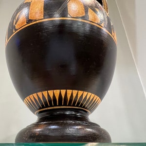 May include: Black and brown ceramic amphora with geometric designs and a black base. The amphora is on a glass shelf.