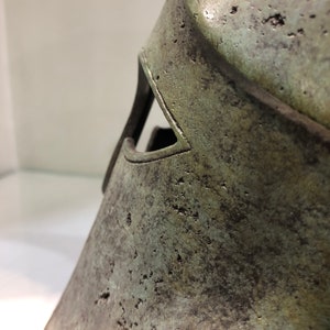 May include: A close-up of a bronze helmet with a green patina. The helmet has a distinctive shape with a raised ridge and a small eye hole.