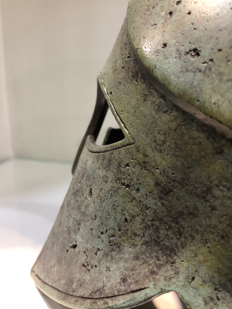 May include: A close-up of a bronze helmet with a green patina. The helmet has a distinctive shape with a raised ridge and a small eye hole.