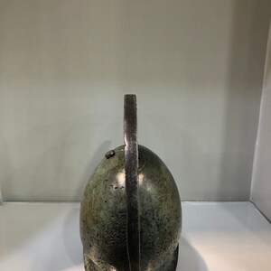 May include: A bronze helmet with a long, curved crest. The helmet is green and has a textured surface.