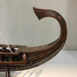 Puede incluir: Un modelo de barco de madera con una proa curva y detallada. El barco es marr&oacute;n y tiene una cuerda blanca atada a la parte delantera.