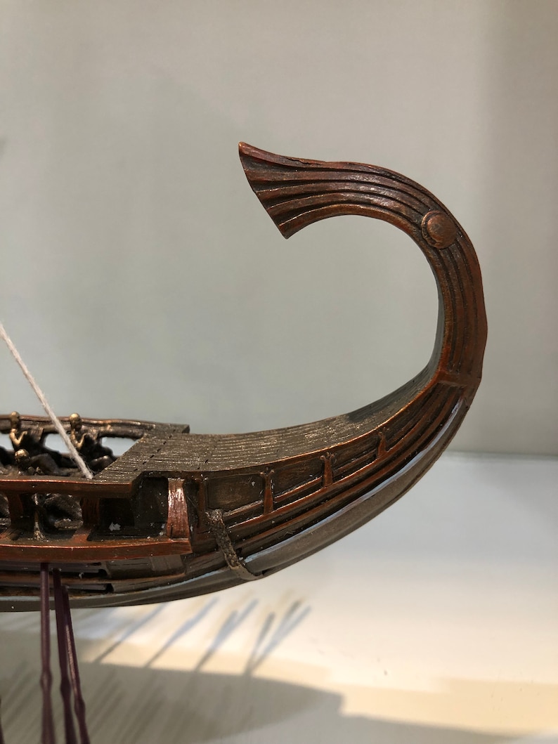 Puede incluir: Un modelo de barco de madera con una proa curva y detallada. El barco es marr&oacute;n y tiene una cuerda blanca atada a la parte delantera.