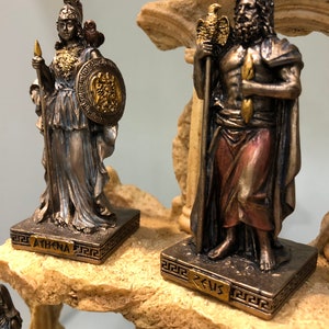May include: A set of two bronze Greek god statues, Athena and Zeus, on a stone base. The statues are detailed and feature gold accents. The base is decorated with a Greek key pattern.