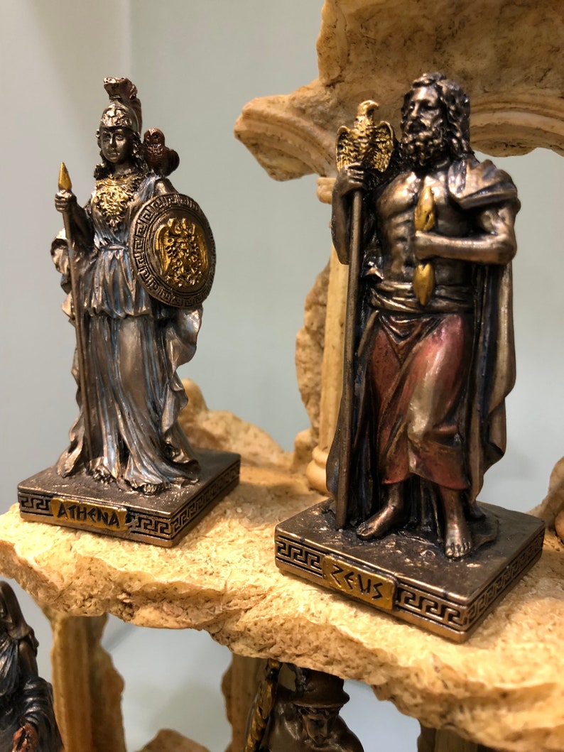 May include: A set of two bronze Greek god statues, Athena and Zeus, on a stone base. The statues are detailed and feature gold accents. The base is decorated with a Greek key pattern.