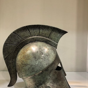 May include: A bronze Greek warrior helmet with a plume and a decorative band around the top. The helmet is weathered and has a green patina.