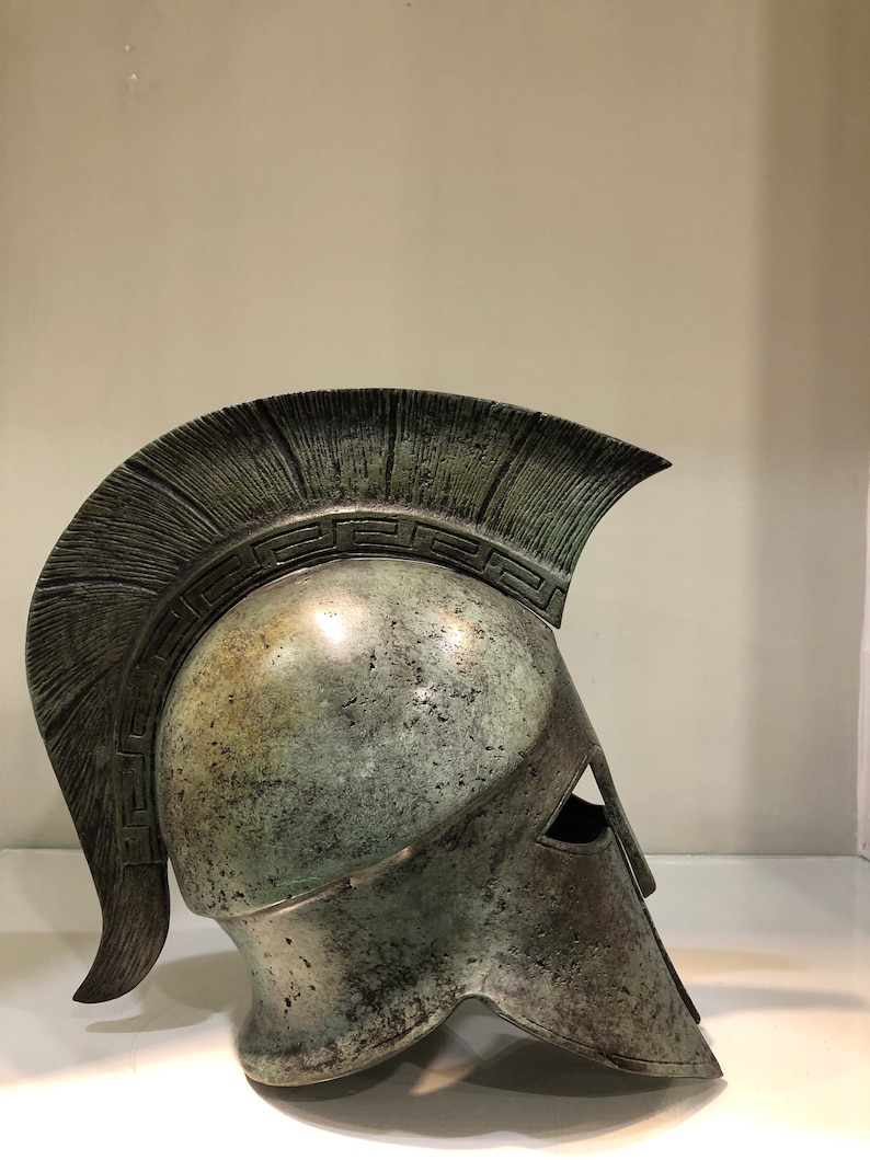 May include: A bronze Greek warrior helmet with a plume and a decorative band around the top. The helmet is weathered and has a green patina.