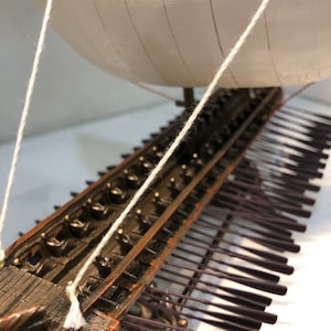 Puede incluir: Un modelo detallado de un barco trirreme griego antiguo de madera con velas blancas y una fila de remos. El barco est&aacute; hecho de metal y tiene un acabado marr&oacute;n oscuro. El modelo est&aacute; sobre una superficie blanca.