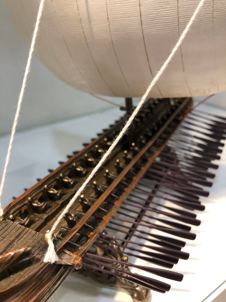 Puede incluir: Un modelo detallado de un barco trirreme griego antiguo de madera con velas blancas y una fila de remos. El barco est&aacute; hecho de metal y tiene un acabado marr&oacute;n oscuro. El modelo est&aacute; sobre una superficie blanca.