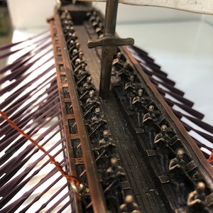 Puede incluir: Un modelo detallado de una galera romana con una vela blanca y un m&aacute;stil de bronce. El barco est&aacute; lleno de filas de figuras en miniatura de bronce de soldados romanos, todos mirando hacia adelante. El casco del barco est&aacute; hecho de madera oscura y tiene un ribete de color cobre.