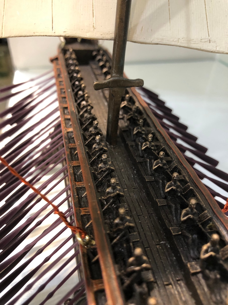 Puede incluir: Un modelo detallado de una galera romana con una vela blanca y un m&aacute;stil de bronce. El barco est&aacute; lleno de filas de figuras en miniatura de bronce de soldados romanos, todos mirando hacia adelante. El casco del barco est&aacute; hecho de madera oscura y tiene un ribete de color cobre.