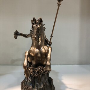 K&ouml;nnte beinhalten: Bronzefarbene Statue einer Figur auf einem Pferd, die aus stilisierten Wellen auftaucht. Die Figur h&auml;lt einen Dreizack und ein kleines Objekt. Die Statue ist detailliert und scheint eine mythologische oder historische Darstellung zu sein.