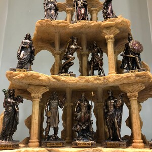 May include: A set of 12 bronze-colored Greek god and goddess figurines arranged on a tiered, rocky, and columned display stand. The figurines are arranged in three tiers, with four figurines on each tier. The figurines are all standing or sitting, and they are all wearing robes or armor. The stand is made of a light brown material that resembles stone.