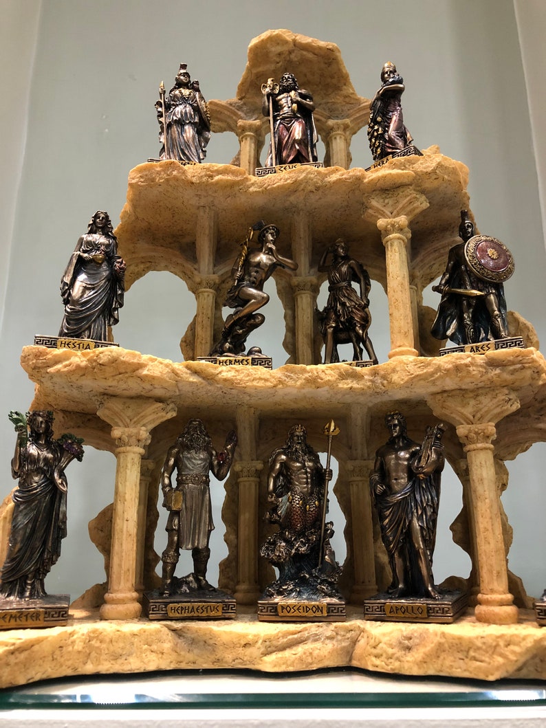 May include: A set of 12 bronze-colored Greek god and goddess figurines arranged on a tiered, rocky, and columned display stand. The figurines are arranged in three tiers, with four figurines on each tier. The figurines are all standing or sitting, and they are all wearing robes or armor. The stand is made of a light brown material that resembles stone.