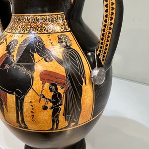 May include: Black and orange ancient Greek amphora with two handles decorated with a black and white scene of a man leading a horse, a man carrying a large object, and a man holding a staff. The amphora has the text "KASTOP", "TYNNA", and "AVVAPOS" written on it.