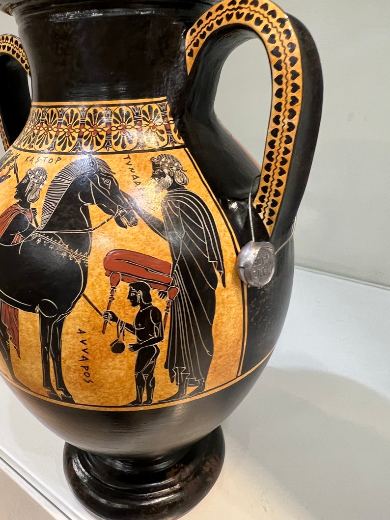 May include: Black and orange ancient Greek amphora with two handles decorated with a black and white scene of a man leading a horse, a man carrying a large object, and a man holding a staff. The amphora has the text "KASTOP", "TYNNA", and "AVVAPOS" written on it.