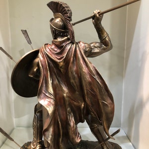 May include: Bronze statue of a Greek warrior holding a spear and shield. The warrior is wearing a helmet with a red plume and a red cape. The statue is standing on a rocky base.
