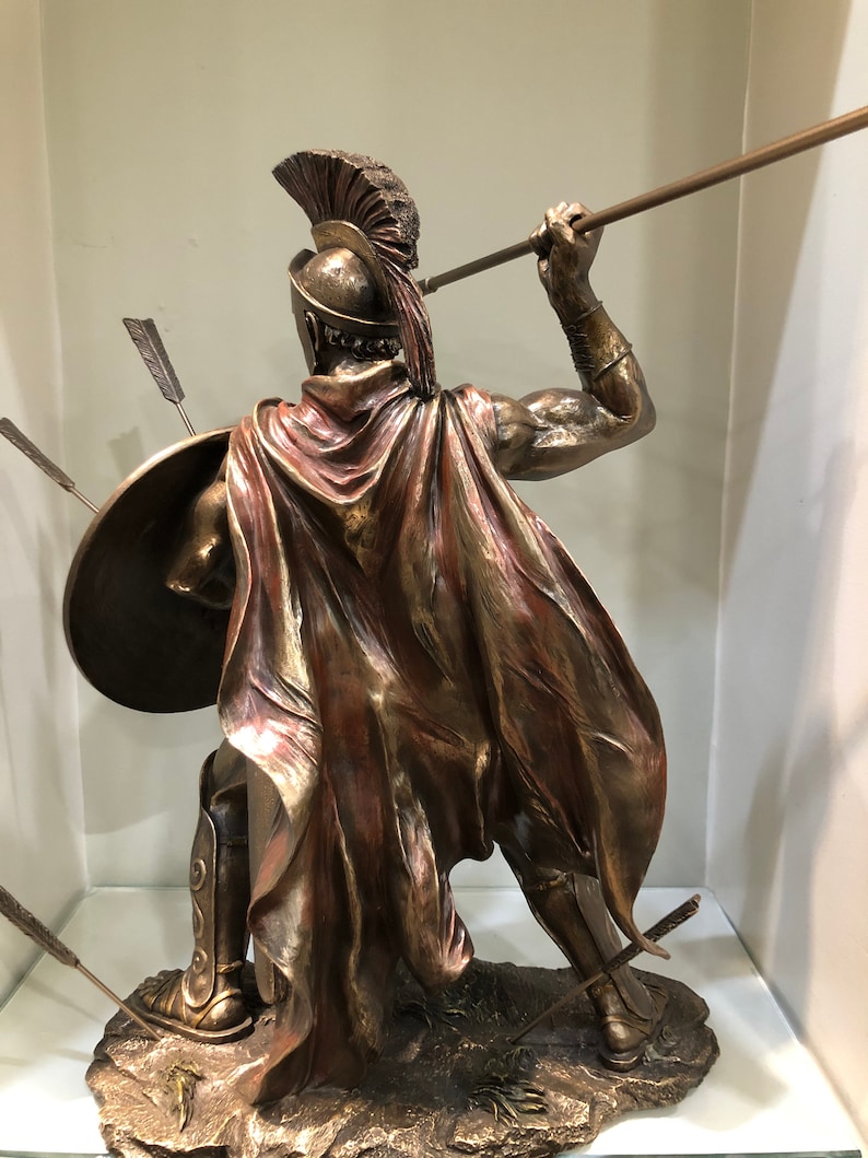 May include: Bronze statue of a Greek warrior holding a spear and shield. The warrior is wearing a helmet with a red plume and a red cape. The statue is standing on a rocky base.
