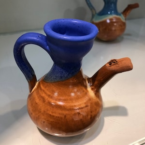 May include: Two small ceramic pitchers with brown and blue glaze. The pitcher on the left has a blue spout and a brown handle. The pitcher on the right has a blue spout and a brown handle.