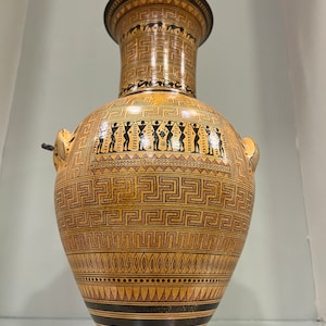Puede incluir: Un jarrón griego antiguo de color marrón y negro con patrones geométricos y figuras. El jarrón tiene un cuello estrecho y un cuerpo ancho. Los patrones incluyen un meandro, triángulos y puntos.