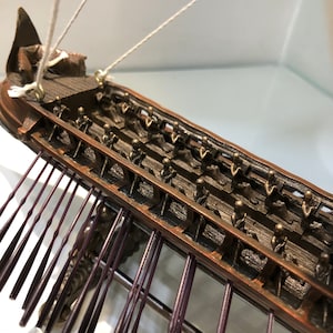 Puede incluir: Un modelo detallado de un antiguo barco de guerra trirreme griego con filas de remos y una tripulaci&oacute;n de figuras en miniatura. El barco est&aacute; hecho de madera y pintado en tonos marrones y negros. Los remos est&aacute;n hechos de metal y pintados de color morado oscuro.