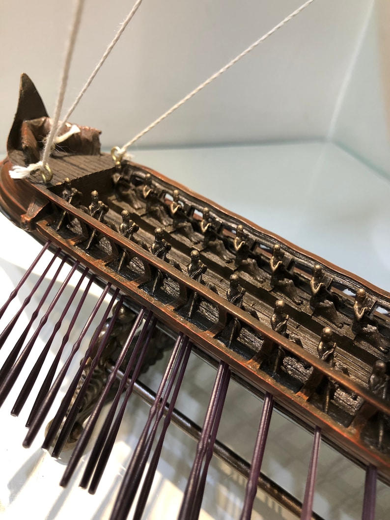 Puede incluir: Un modelo detallado de un antiguo barco de guerra trirreme griego con filas de remos y una tripulaci&oacute;n de figuras en miniatura. El barco est&aacute; hecho de madera y pintado en tonos marrones y negros. Los remos est&aacute;n hechos de metal y pintados de color morado oscuro.