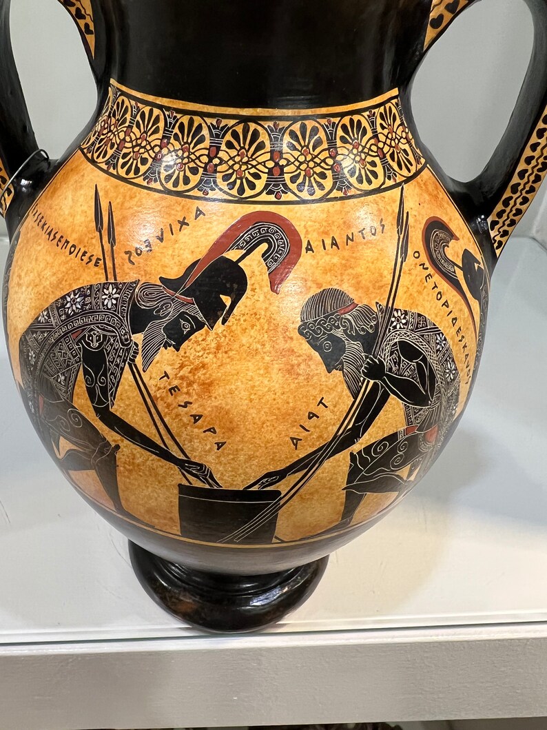 May include: Black and brown ceramic amphora with a black and gold geometric design around the top. The amphora features a scene of two figures in black silhouette with spears and a cauldron. The figures are labeled in Greek script: "TESAPA", "AIANT", "ROUVIKA", "AIANTOS", "ONETOPIESE".