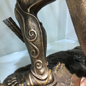 May include: Bronze statue of a warrior's leg wearing a detailed, ornate, and intricately designed shin guard with a decorative pattern of swirls. The warrior's foot is wearing a sandal with straps.