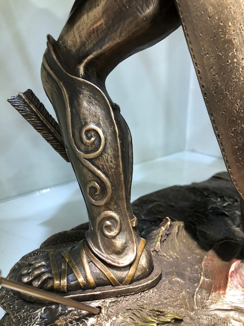 May include: Bronze statue of a warrior's leg wearing a detailed, ornate, and intricately designed shin guard with a decorative pattern of swirls. The warrior's foot is wearing a sandal with straps.