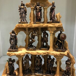 May include: A set of 12 bronze-colored Greek god and goddess figurines arranged on a stone-like display stand. The figurines are arranged in three rows of four. Each figurine is standing on a small base with the name of the god or goddess inscribed on it. The names are: Hestia, Athena, Hera, Hermes, Artemis, Ares, Demeter, Hephaestus, Poseidon, Apollo, Aphrodite, and Artemis.