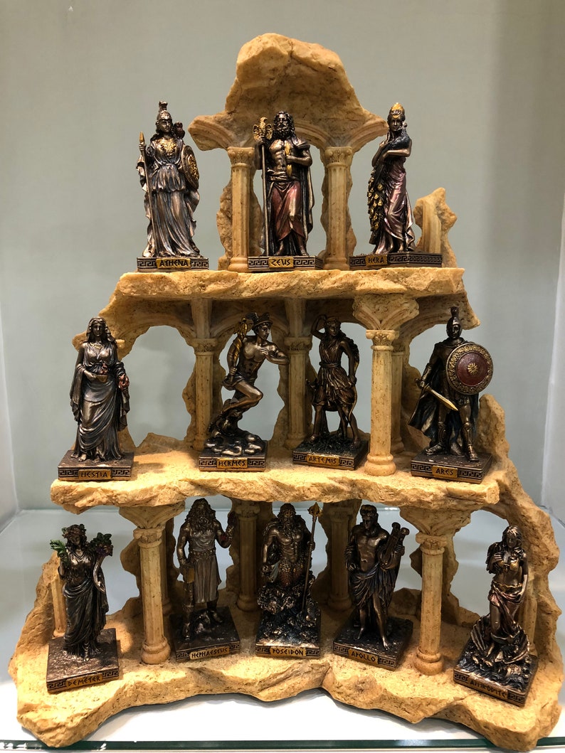 May include: A set of 12 bronze-colored Greek god and goddess figurines arranged on a stone-like display stand. The figurines are arranged in three rows of four. Each figurine is standing on a small base with the name of the god or goddess inscribed on it. The names are: Hestia, Athena, Hera, Hermes, Artemis, Ares, Demeter, Hephaestus, Poseidon, Apollo, Aphrodite, and Artemis.