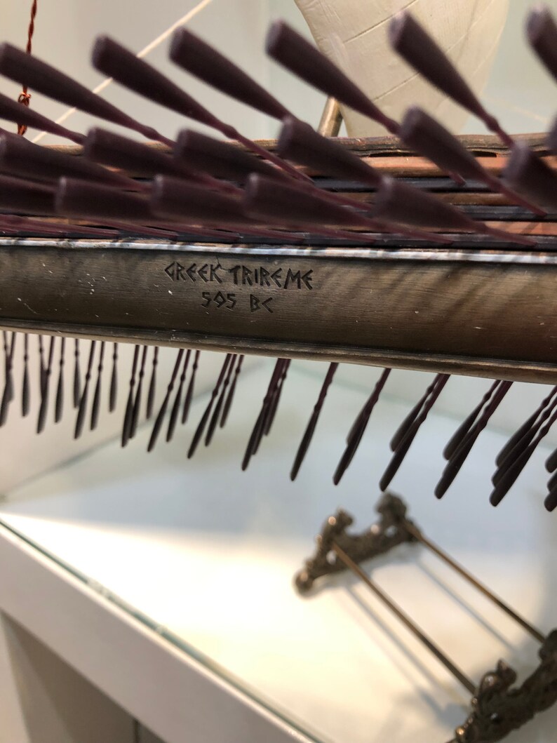 Puede incluir: Primer plano de una maqueta de una trirreme griega, un tipo de barco de guerra griego antiguo, con la inscripci&oacute;n "GREEK TRIREME 505 BC" en el lateral.