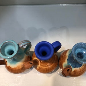 May include: Three small ceramic pitchers with brown, blue, and light blue glaze. The pitchers have a rounded body and a spout on the side. The pitchers are arranged in a row on a white surface.