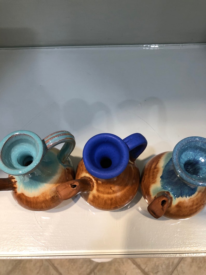 May include: Three small ceramic pitchers with brown, blue, and light blue glaze. The pitchers have a rounded body and a spout on the side. The pitchers are arranged in a row on a white surface.