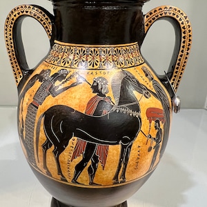 May include: Black and red Greek amphora with two handles and a black and white painted scene of a man holding a horse's reins. The scene includes the Greek words "KASTOR", "TYNAEOS", "DIOSKOUROI", and "ANVAOS".