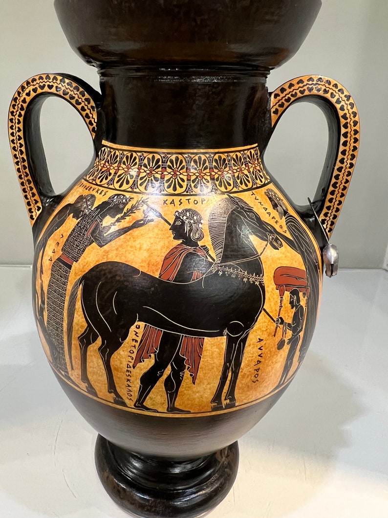May include: Black and red Greek amphora with two handles and a black and white painted scene of a man holding a horse's reins. The scene includes the Greek words "KASTOR", "TYNAEOS", "DIOSKOUROI", and "ANVAOS".
