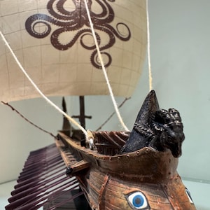 May include: A model ship featuring a brown wooden hull with painted blue eyes on the bow, a black ram figurehead, and a white sail printed with a brown octopus graphic. The vessel includes multiple dark oars extending from the sides and white rigging.