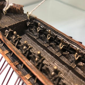 Puede incluir: Primer plano del interior de un modelo de barco de madera. La cubierta del barco est&aacute; hecha de madera oscura y tiene filas de peque&ntilde;as figuras detalladas de personas sentadas en bancos. Las figuras est&aacute;n hechas de un material de color bronce.