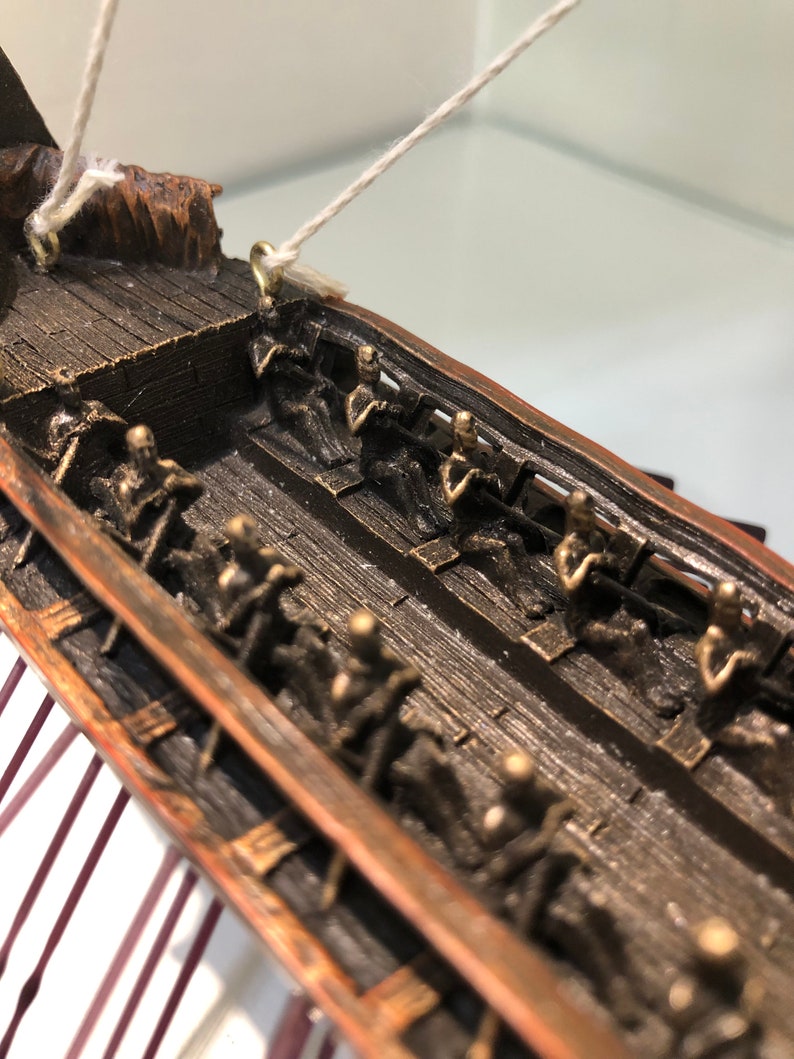 Puede incluir: Primer plano del interior de un modelo de barco de madera. La cubierta del barco est&aacute; hecha de madera oscura y tiene filas de peque&ntilde;as figuras detalladas de personas sentadas en bancos. Las figuras est&aacute;n hechas de un material de color bronce.