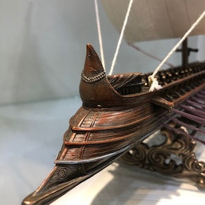 Puede incluir: Un modelo detallado de un barco de madera con una vela blanca, un soporte decorativo y un acabado en bronce. El barco tiene una proa puntiaguda y una fila de remos.