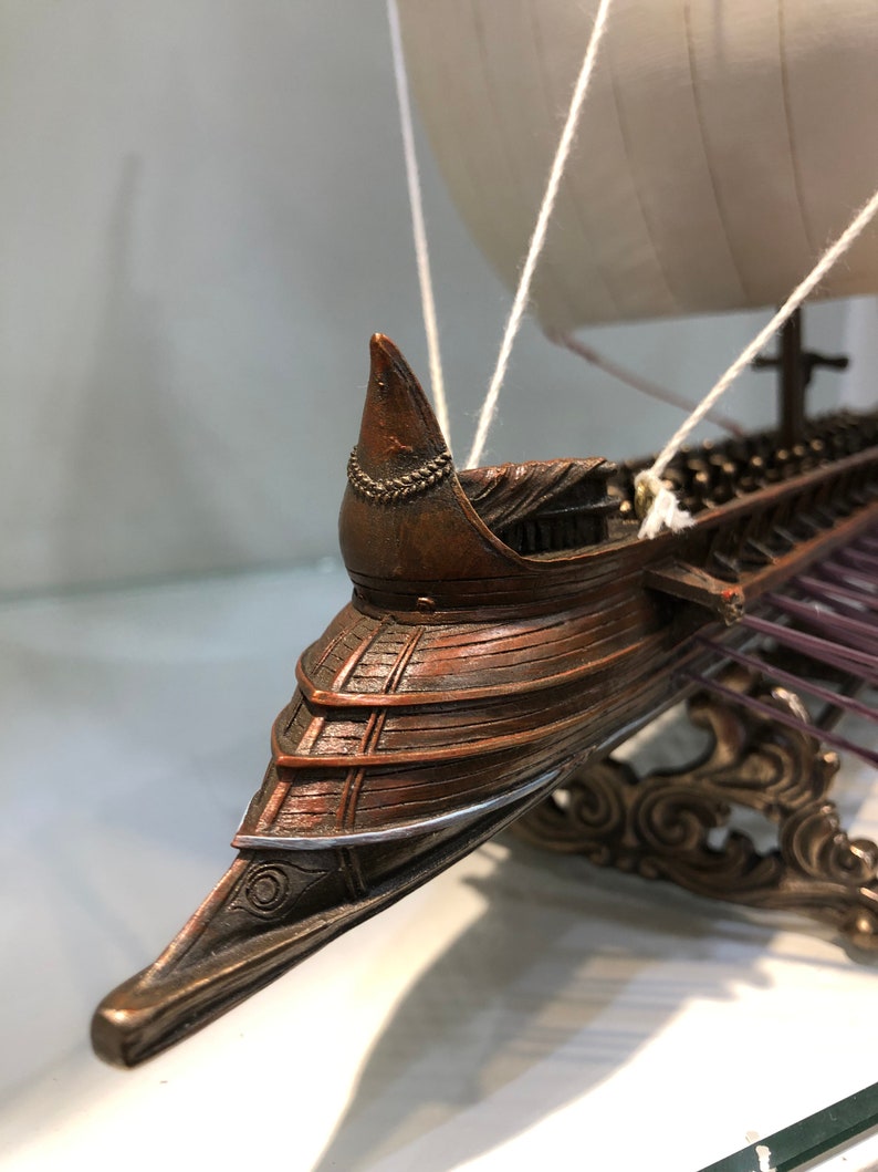 Puede incluir: Un modelo detallado de un barco de madera con una vela blanca, un soporte decorativo y un acabado en bronce. El barco tiene una proa puntiaguda y una fila de remos.