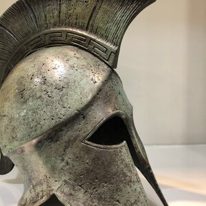 May include: A bronze Greek warrior helmet with a crested top and a face mask. The helmet has a decorative pattern around the top.