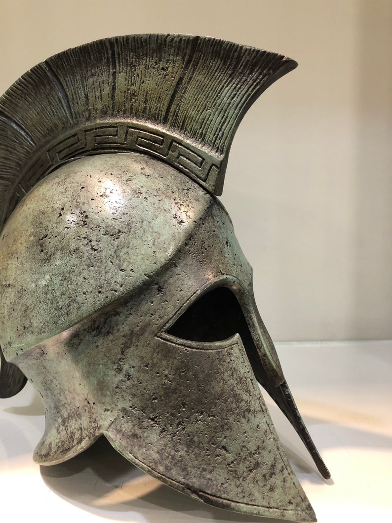 May include: A bronze Greek warrior helmet with a crested top and a face mask. The helmet has a decorative pattern around the top.
