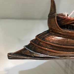 Puede incluir: Primer plano de la proa de un modelo de barco de madera. La proa est&aacute; pintada en color bronce y tiene un dise&ntilde;o de ojo decorativo. La proa del barco es puntiaguda y tiene una aleta decorativa.