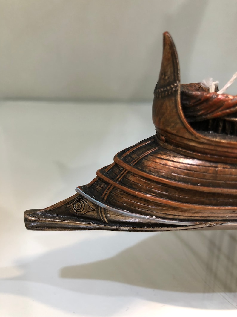 Puede incluir: Primer plano de la proa de un modelo de barco de madera. La proa est&aacute; pintada en color bronce y tiene un dise&ntilde;o de ojo decorativo. La proa del barco es puntiaguda y tiene una aleta decorativa.