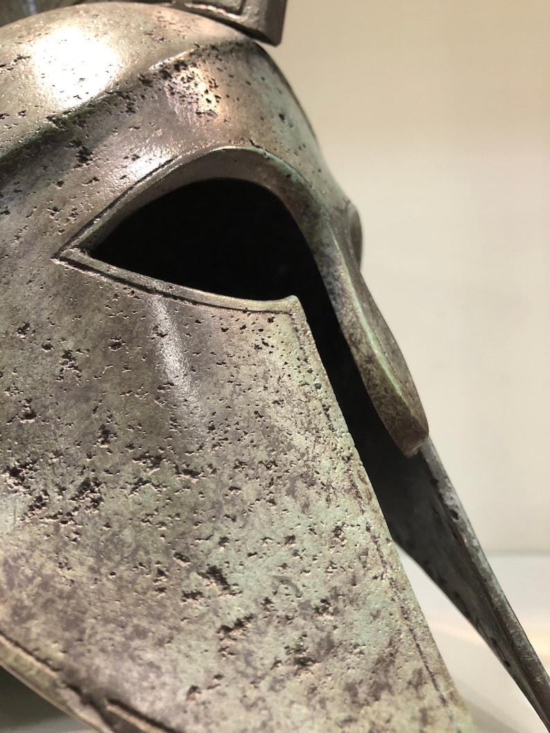 May include: A bronze Greek warrior helmet with a weathered patina. The helmet has a distinctive pointed crest and a face mask with a single eye opening.