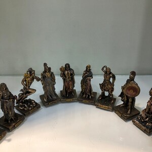 May include: A set of ten small, gold-toned figurines of Greek gods and goddesses. Each figurine is standing on a square base with the name of the god or goddess written on it. The figurines are arranged in a semi-circle on a white surface.