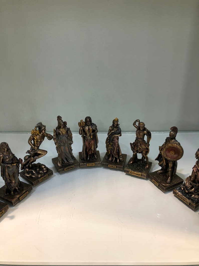May include: A set of ten small, gold-toned figurines of Greek gods and goddesses. Each figurine is standing on a square base with the name of the god or goddess written on it. The figurines are arranged in a semi-circle on a white surface.
