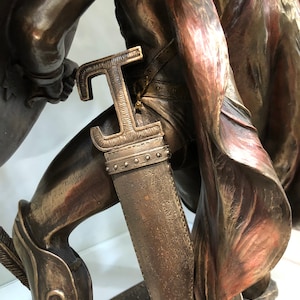 May include: Bronze statue of a warrior holding a sword and shield. The warrior is wearing a red cape and has a detailed, textured surface.